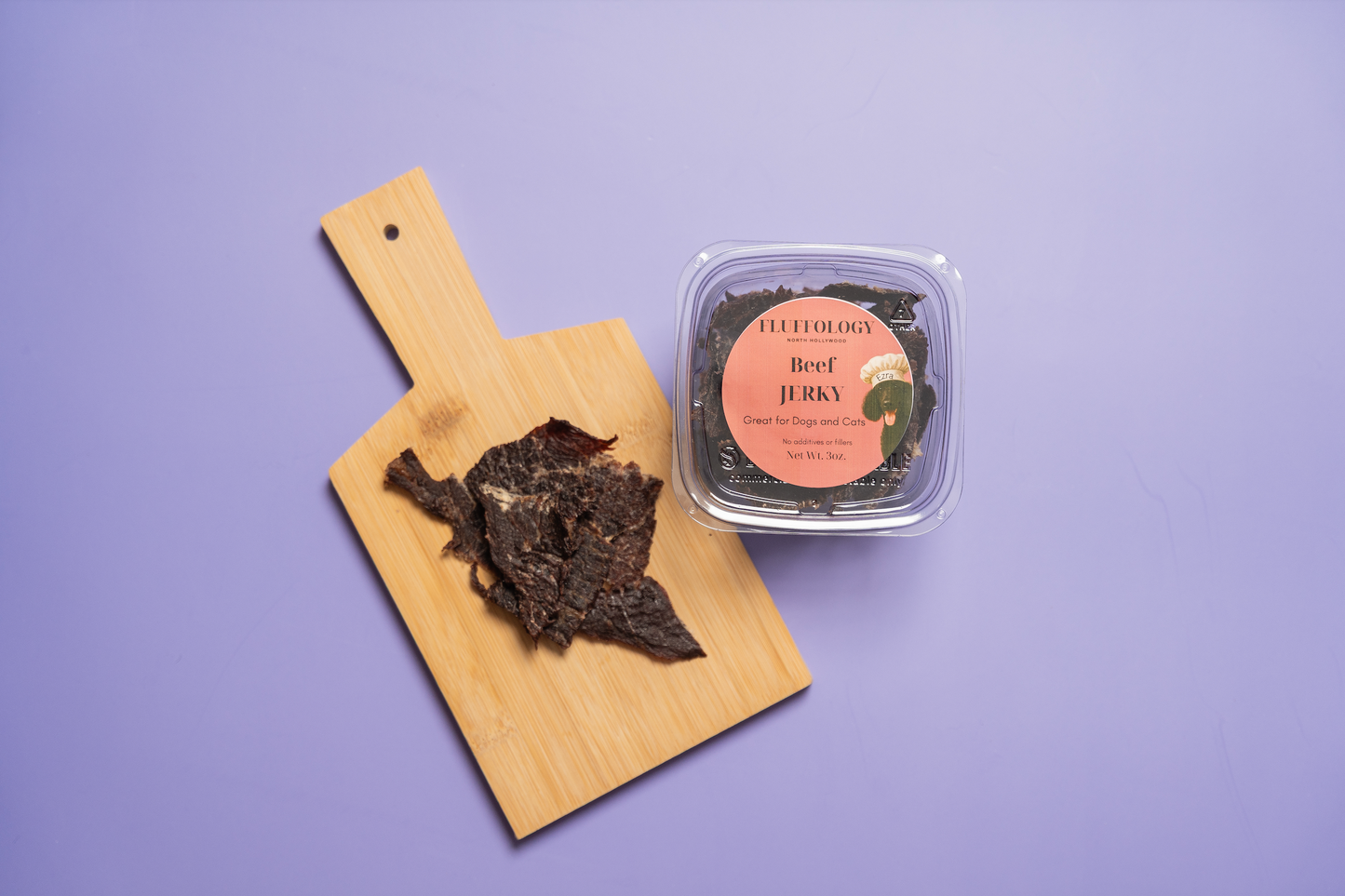 Beef jerky on a wooden paddle with a container of beef jerky on a purple background. Fluffology Dehydrated Beef Jerky