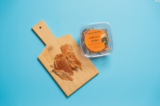 Container of chicken jerky on a wooden board with a blue background. Fluffology Dehydrated Chicken Jerky
