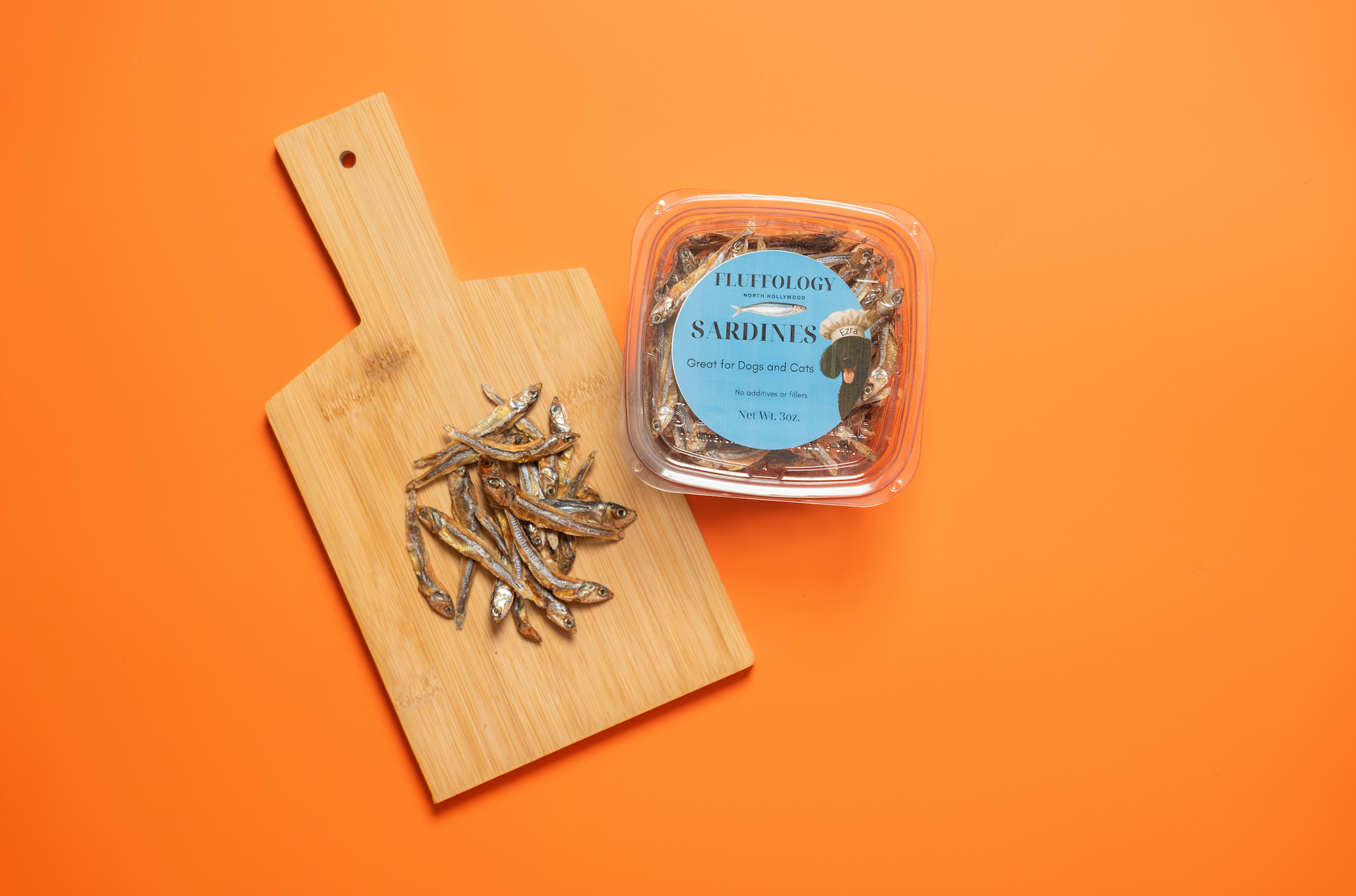 Dried sardines on a wooden cutting board with a container on an orange background. Fluffology Dehydrated Sardines